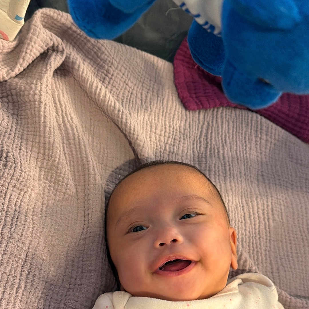 Danielle Anaia is registered to the contest to win money with this photo: baby, infant, child, smiling, happy, portrait, lying_down, blanket, quilt, stuffed_animal, plush_toy, blue_toy, onesie, home, indoor, cozy, bed, bedding, face, photography
