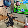 Ayaz is registered to the contest to win money with this photo: toddler, child, balloons, living_room, tv, cartoon, playmat, striped_onesie, reaching, smile, toys, cabinet, wood_floor, celebration, balloon_strings, indoor, television_subtitles, happy, sitting, playtime