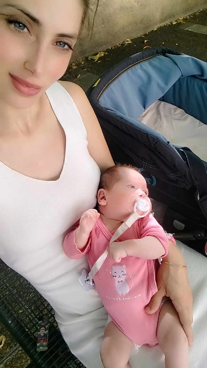 Mila participe au concours pour gagner de l'argent avec cette photo : woman, baby, pacifier, pink_onesie, cat_illustration, bench, stroller, outdoor, child, infant, holding, white_tank_top, casual_clothing, skin, face, hand, nature, daylight, parent, cute