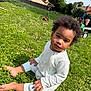 toddler, child, grass, outdoor, sunny, curly_hair, white_onesie, barefoot, greenery, flowers, backyard, toy_scooter, person, nature, summer, playtime, sitting, casual, daylight, portrait