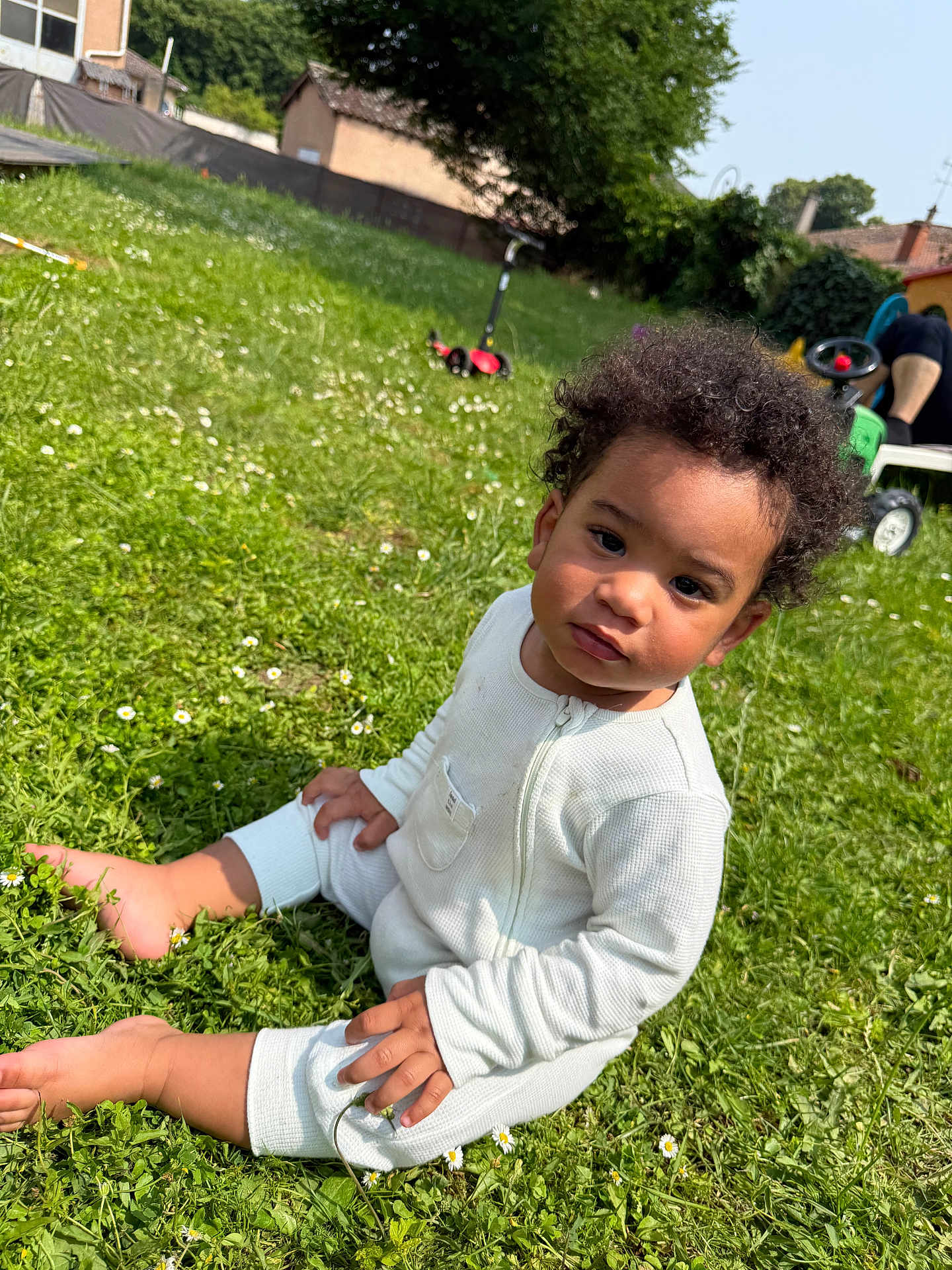 Harlow participe au concours pour gagner de l'argent avec cette photo : toddler, child, grass, outdoor, sunny, curly_hair, white_onesie, barefoot, greenery, flowers, backyard, toy_scooter, person, nature, summer, playtime, sitting, casual, daylight, portrait