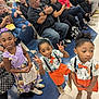 children, kids, boys, girl, certificate, event, crowd, seated_adults, clothing, orange_shorts, bow_tie, peace_sign, floor, chairs, indoor, people, smiling, hair_bow, shoes, casual
