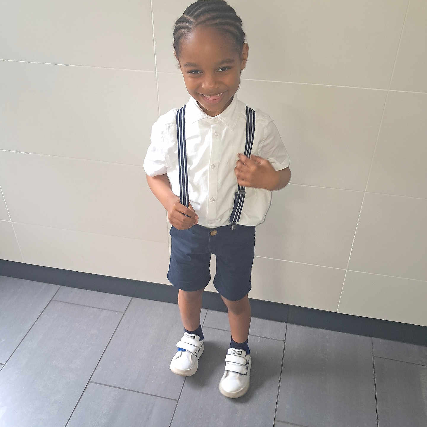 Jemmy a rejoint le concours — aidez-le/la à gagner de superbes lots ! boy, braided_hair, casual_clothing, child, cute, fashion, floor_tiles, footwear, happy, indoors, person, portrait, shorts, smile, sneakers, standing, suspenders, wall, white_shirt, young