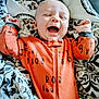 baby, infant, laughing, orange_onesie, blanket, cozy, happy, smiling, child, cute, face, hands, lying_down, patterned_blanket, indoor, portrait, young_child, soft_texture, comfortable, expressive