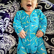 Remington is registered to the contest to win money with this photo: baby, infant, blue_onesie, patterned_blanket, smiling, happy, child, face, person, couch, soft_fabric, indoors, cute, young_child, joyful, relaxed, sleepwear, colorful, cozy, comfort