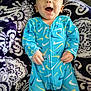 baby, infant, blue_onesie, patterned_blanket, smiling, happy, child, face, person, couch, soft_fabric, indoors, cute, young_child, joyful, relaxed, sleepwear, colorful, cozy, comfort