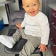 Lyzandro a rejoint le concours — aidez-le/la à gagner de superbes lots ! toddler, child, smiling, white_sweater, gray_chair, white_sneakers, blue_eyes, indoors, kitchen, tile_floor, pants, modern_furniture, happy, sitting, person, cute, young_child, casual_clothing, hand_on_chair, natural_light