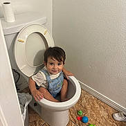 Santiago is registered to the contest to win money with this photo: child, toddler, smiling, bathroom, toilet, toilet_paper, overalls, shirt, sitting, portrait, face, hair, indoor, floor_tiles, wall, toys, colorful, domestic_scene, happy, photography