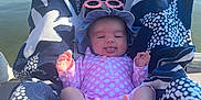 Layla is registered to the contest to win money with this photo: baby, infant, towel, pink_suit, hat, sunglasses, lake, water, outdoor, sunny, feet, tongue_out, smile, relaxing, summer, child, person, seat, blue, white