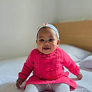 Aintsoa a rejoint le concours — aidez-le/la à gagner de superbes lots ! baby, child, smiling, sitting, pink_sweater, white_tights, headband, flower, indoors, bed, blanket, knitwear, portrait, cute, happy, infant, person, clothing, soft_light, home
