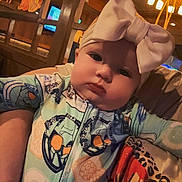 Zoey joined the competition — help win amazing prizes! baby, child, headband, bow, onesie, indoor, warm_lighting, person, face, expression, holding, patterned_clothing, lamp, wooden_furniture, cozy, curious, infant, portrait, candid, closeup