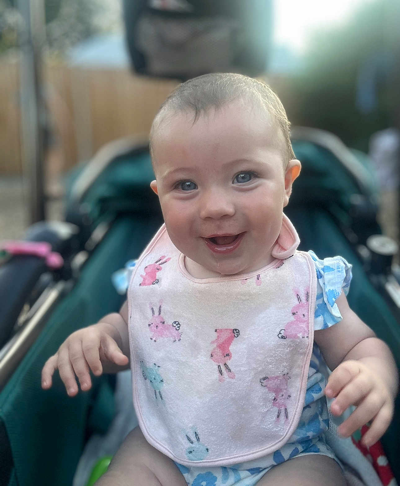 Skylah is registered to the contest to win money with this photo: baby, smiling, bib, bunny_pattern, blue_outfit, stroller, happy, child, infant, face, hands, outdoor, daylight, cute, portrait, clothing, person, skin, head, eyes