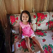 Beatrace is registered to the contest to win money with this photo: child, girl, smiling, pink_clothing, couch, floral_pattern, indoor, curtains, happy, sitting, barefoot, home, furniture, portrait, cute, young_child, person, casual, daylight, relaxed