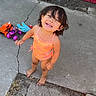 toddler, child, smiling, orange_swimsuit, barefoot, toys, toy_trucks, plush_dinosaur, outdoor, concrete, cracks, broom, happy, curly_hair, sunny, playtime, person, standing, cute, daylight