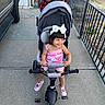 toddler, child, tricycle, stroller, outdoor, sidewalk, smile, happy, person, footwear, clothing, hand, fence, vehicle, concrete, hair_bow, shorts, sunlight, play, family
