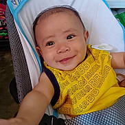 Jeron joined the competition — help win amazing prizes! baby, child, smiling, yellow_bib, seat, indoor, person, happy, infant, reaching, clothing, face, cute, baby_seat, arm, closeup, head, expression, fabric, background