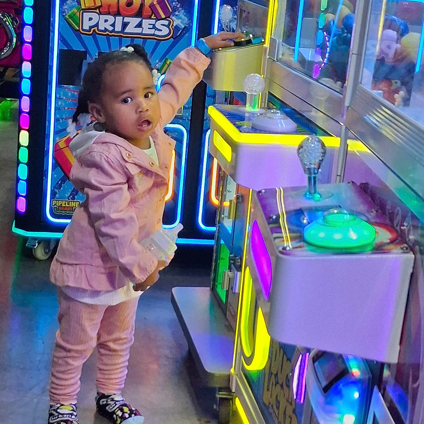 Akayla is registered to the contest to win money with this photo: arcade, arcade_machine, braided_hair, bright_lighting, child, claw_machine, colorful, electronic_game, entertainment_center, game_room, indoor, looking_at_camera, neon_lights, pink_outfit, portrait, prize_machine, reaching, sneakers, standing, toddler