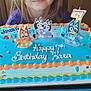 child, girl, birthday_cake, cake, candle, number_7, cartoon_characters, blue_frosting, birthday, smiling, face, celebration, table, chair, indoor, party, happy, icing, decorations, young