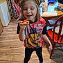 Kara is registered to the contest to win money with this photo: child, tiger_ears, smiling, indoor, kitchen, wood_floor, table, chair, socks, food, happy, person, curtains, window, cup, toy, shirt, black_pants, red_chair, snack