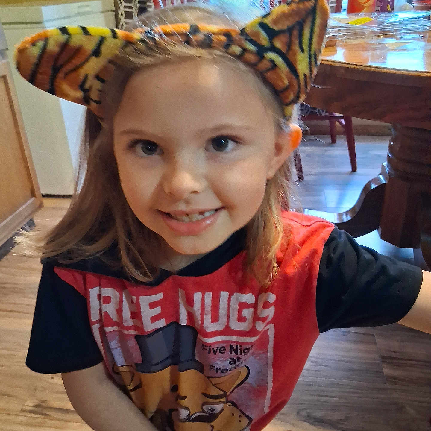 Kara is registered to the contest to win money with this photo: casual, chair, child, curtain, cute, dining_table, graphic_tee, happy, headband, home, indoor, kitchen, person, portrait, smiling, tiger_ears, tshirt, window, wooden_floor, young