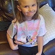 Kara is registered to the contest to win money with this photo: child, girl, bow, remote_control, tshirt, bluey, couch, leather, smile, hair, sitting, indoor, floor, shoe, pillow, wood_floor, casual_clothing, person, happy, home