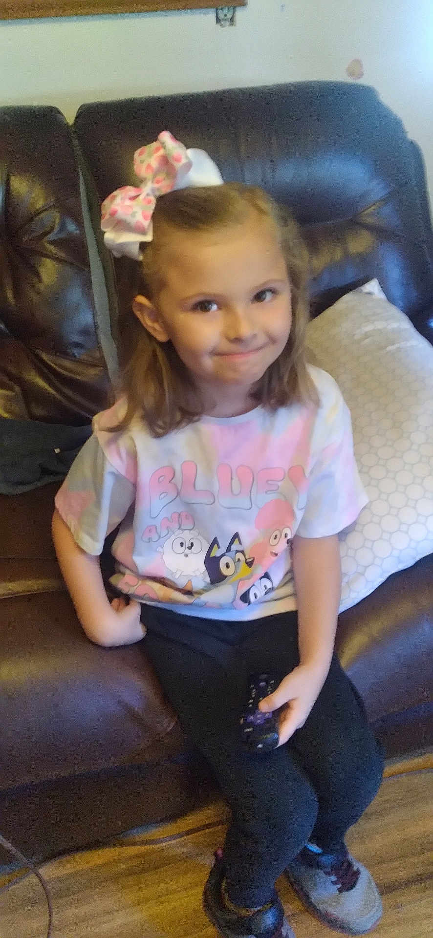 Kara is registered to the contest to win money with this photo: child, girl, bow, remote_control, tshirt, bluey, couch, leather, smile, hair, sitting, indoor, floor, shoe, pillow, wood_floor, casual_clothing, person, happy, home