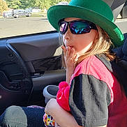 Kara joined the competition — help win amazing prizes! child, hat, sunglasses, car_seat, plush_toy, window, sunlight, seatbelt, car_interior, person, casual_clothing, red_shirt, black_sleeves, leggings, colorful_toy, daylight, trees, outdoor_view, reflection, seat