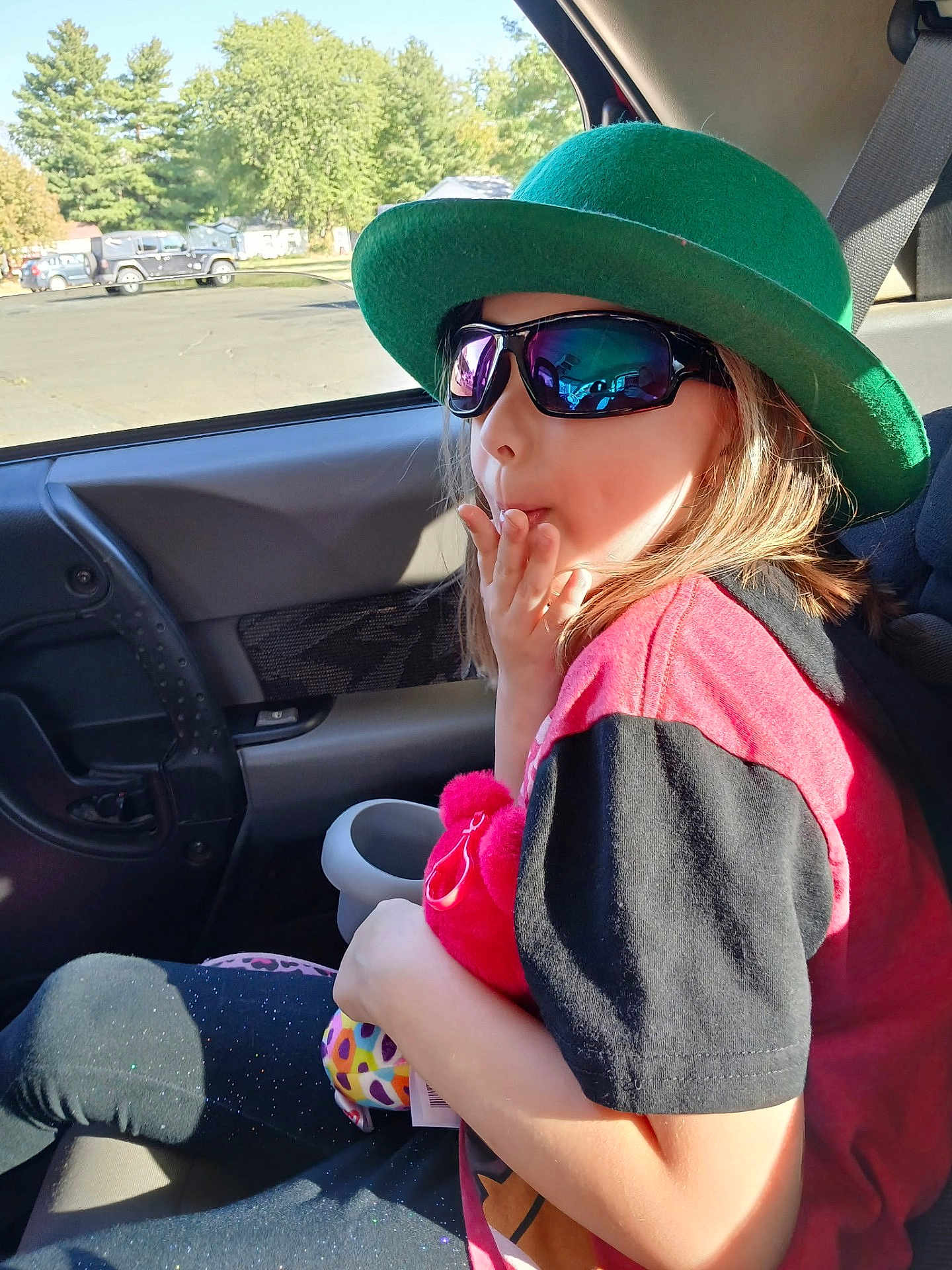 Kara joined the competition — help win amazing prizes! child, hat, sunglasses, car_seat, plush_toy, window, sunlight, seatbelt, car_interior, person, casual_clothing, red_shirt, black_sleeves, leggings, colorful_toy, daylight, trees, outdoor_view, reflection, seat