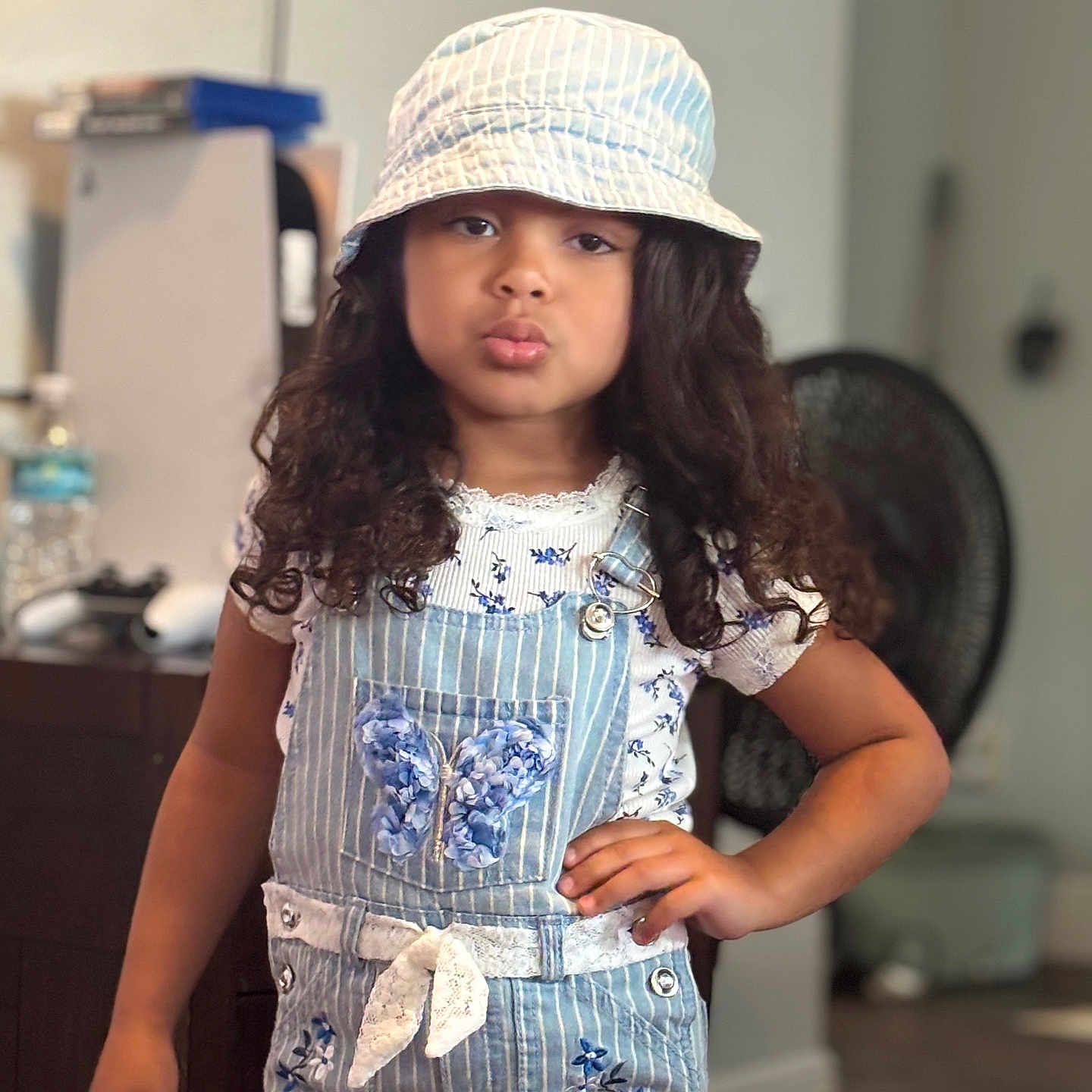 Deliyah is registered to the contest to win money with this photo: blue, blurred_background, bucket_hat, butterfly_patch, casual_clothing, child, curly_hair, cute, fashion, floral_pattern, furniture, girl, hand_on_hip, indoor, overalls, portrait, pose, pout, short_sleeves, young