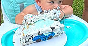 Kohen is registered to the contest to win money with this photo: baby, birthday_cake, high_chair, party_hat, frosting, messy, outdoor, grass, celebration, child, blue, racecar, sign, half_birthday, food, smile, person, sitting, cute, festive