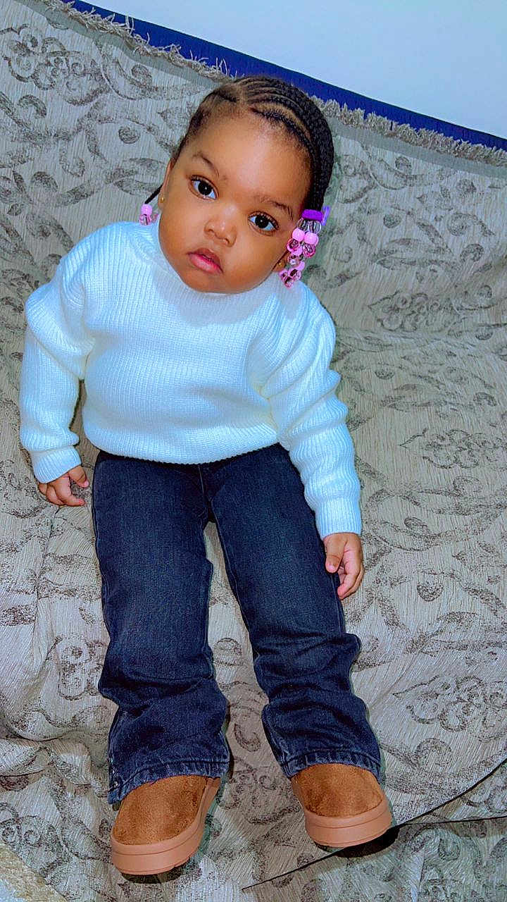 Lya participe au concours pour gagner de l'argent avec cette photo : child, toddler, braided_hair, pink_beads, white_sweater, jeans, boots, couch, patterned_couch, indoor, sitting, curious_expression, young_child, portrait, face, person, casual_clothing, home, furniture, cute