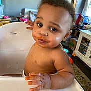 Demitruis J joined the competition — help win amazing prizes! toddler, child, high_chair, messy_face, food, indoor, curly_hair, brown_skin, looking_at_camera, hand, furniture, toy, room, window, curtain, cabinet, carpet, table, expression, cute