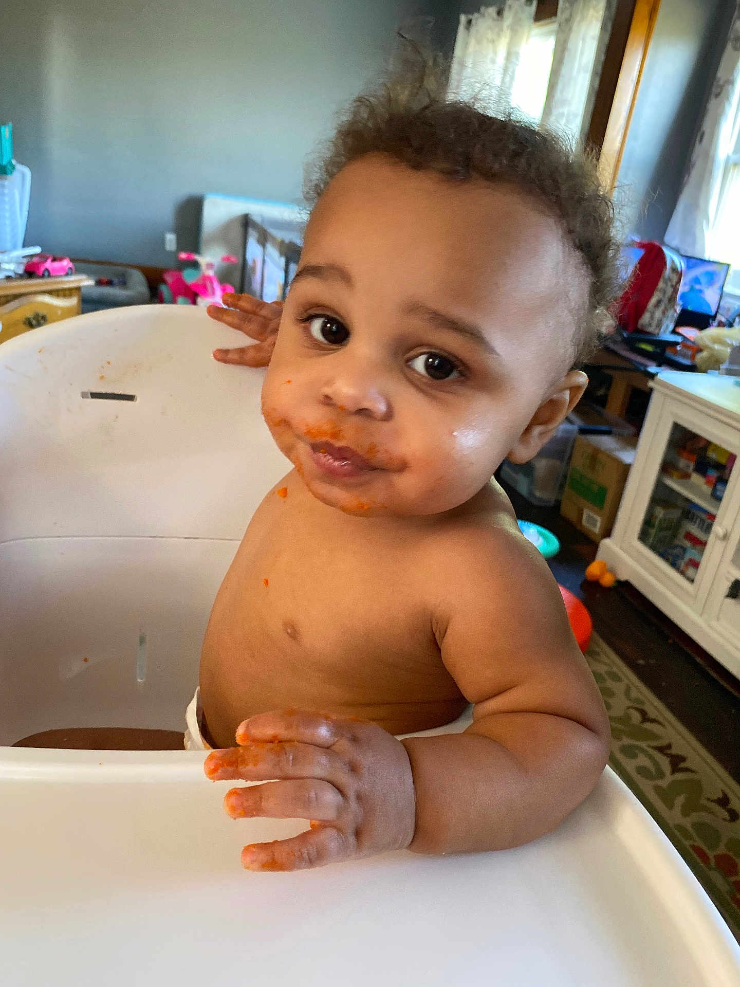 Demitruis J joined the competition — help win amazing prizes! toddler, child, high_chair, messy_face, food, indoor, curly_hair, brown_skin, looking_at_camera, hand, furniture, toy, room, window, curtain, cabinet, carpet, table, expression, cute