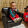toddler, child, smile, chair, black_leather, red_pants, green_shirt, festive_clothing, toy_car, indoor, tile_floor, bamboo_screen, happy, sitting, footwear, playful, thumbs_up, baby, person, cute