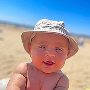 Issa a rejoint le concours — aidez-le/la à gagner de superbes lots ! baby, child, hat, bucket_hat, smiling, beach, sand, sunny, outdoor, arm, skin, face, headwear, summer, portrait, infant, blue_sky, sunlight, closeup, happy