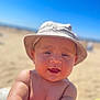 baby, child, hat, bucket_hat, smiling, beach, sand, sunny, outdoor, arm, skin, face, headwear, summer, portrait, infant, blue_sky, sunlight, closeup, happy