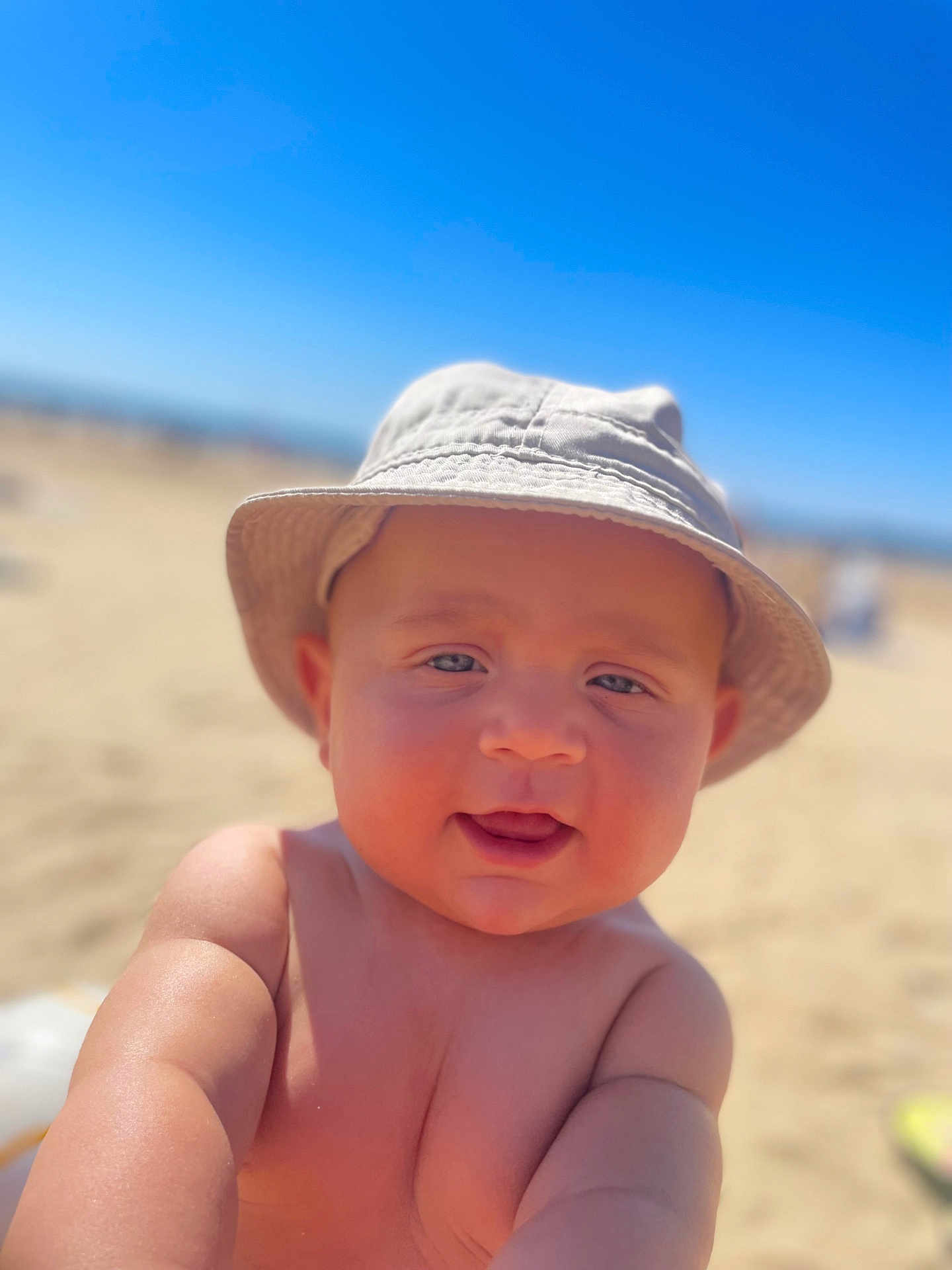 Issa a rejoint le concours — aidez-le/la à gagner de superbes lots ! baby, child, hat, bucket_hat, smiling, beach, sand, sunny, outdoor, arm, skin, face, headwear, summer, portrait, infant, blue_sky, sunlight, closeup, happy