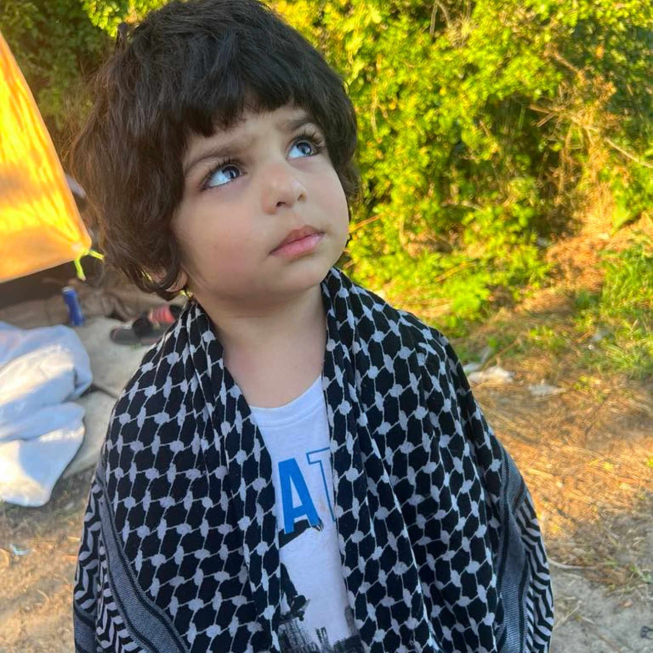 Yousif joined the competition — help win amazing prizes! casual, child, clothing, curious, curly_hair, daylight, expression, greenery, head_and_shoulders, looking_up, nature, outdoor, person, portrait, scarf, standing, sunlight, tent, thoughtful, young_child