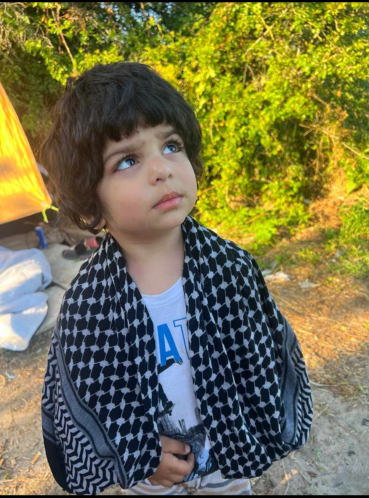 Yousif joined the competition — help win amazing prizes! child, outdoor, greenery, scarf, curly_hair, thoughtful, sunlight, tent, nature, portrait, young_child, clothing, person, daylight, casual, expression, standing, looking_up, curious, head_and_shoulders