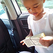 Zylus is registered to the contest to win money with this photo: car_interior, cash, child, counting, curious, daylight, dollar_bills, hands, indoors, money, person, rubber_band, seat, standing, sunlight, vehicle, white_shirt, window, young_child, young_person