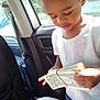 car_interior, cash, child, counting, curious, daylight, dollar_bills, hands, indoors, money, person, rubber_band, seat, standing, sunlight, vehicle, white_shirt, window, young_child, young_person
