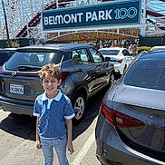 Mohammed is registered to the contest to win money with this photo: child, boy, smiling, parking_lot, car, automobile, roller_coaster, amusement_park, sign, sunny, daytime, casual_clothing, jeans, sneakers, blue_shirt, license_plate, outdoor, metal_structure, people, recreation