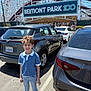 child, boy, smiling, parking_lot, car, automobile, roller_coaster, amusement_park, sign, sunny, daytime, casual_clothing, jeans, sneakers, blue_shirt, license_plate, outdoor, metal_structure, people, recreation