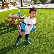Taha joined the competition — help win amazing prizes! child, boy, playground, grass, outdoor, sunlight, picnic_table, tricycle, park, casual_clothing, blue_shoes, white_shirt, happy, active, daytime, trees, fence, play_equipment, water_bottle, recreation
