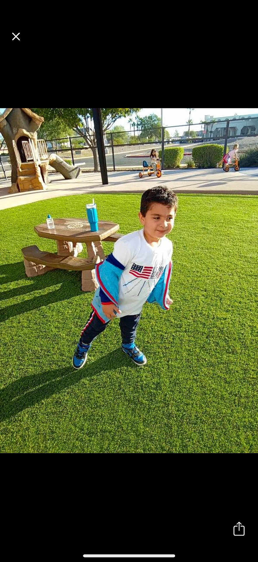 Taha joined the competition — help win amazing prizes! child, boy, playground, grass, outdoor, sunlight, picnic_table, tricycle, park, casual_clothing, blue_shoes, white_shirt, happy, active, daytime, trees, fence, play_equipment, water_bottle, recreation