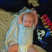 Tommie is registered to the contest to win money with this photo: arms_raised, baby, blanket, blue_onesie, casual, child, clothing, comfort, couch, cute, diaper, face, ghost_face, graphic_tshirt, home_interior, infant, person, relaxed, smiling, soft_lighting