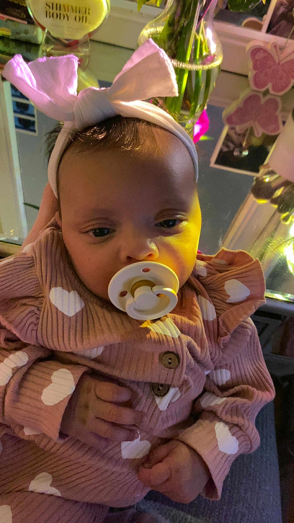 Arielle joined the competition — help win amazing prizes! baby, infant, pacifier, headband, bow, pink_clothing, heart_pattern, onesie, hand, close_up, portrait, indoors, vase, flowers, mirror, reflection, bottle, soft_lighting, sleepy, butterfly_decor