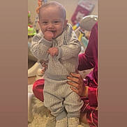 Lorenzo is registered to the contest to win money with this photo: baby, blurred_background, carpet, child, clothing, cute, face, fingers_in_mouth, footwear, hand, happy, home, indoor, infant, pajamas, patterned_clothing, person, smile, support, toy