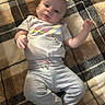 baby, infant, blanket, plaid, pattern, floor, wooden_floor, clothing, grey_pants, tshirt, person, child, smiling, cute, lying_down, indoors, skin, head, hand, foot