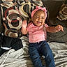 baby, child, smiling, pink_headband, pink_shirt, jeans, white_socks, couch, pillow, blanket, happy, indoors, infant, person, sitting, cute, home, cozy, patterned_pillow, waving